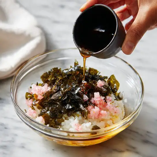 A clear glass bowl contains three main layers: at the bottom, a round mound of white steamed rice; above it, many small pink cubes scattered around the rice; on top, uneven pieces of dark green crispy seaweed spread over the rice and pink cubes. A woman's hand pours a dark brown liquid from a small black cup, creating a thin stream that lands in the bowl. The bowl sits on a white marbled surface with a white cloth in the background. Photo taken with an iphone --ar 4:5 --v 7