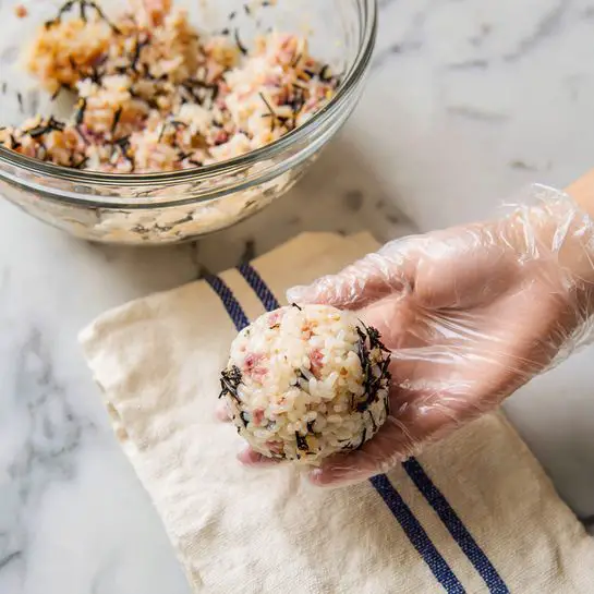 Seven round rice balls are placed on a white scalloped plate, each ball showing a mix of cooked rice, small pinkish cubes of meat, black seaweed pieces, and sesame seeds, topped with a thin line of white sauce. The rice balls have a slightly glossy texture and a mix of light brown and darker bits from the seaweed and sesame. The plate sits on a white marbled surface with a white cloth featuring blue stripes loosely spread behind it. Some sesame seeds are scattered on the surface around the plate. photo taken with an iphone --ar 4:5 --v 7