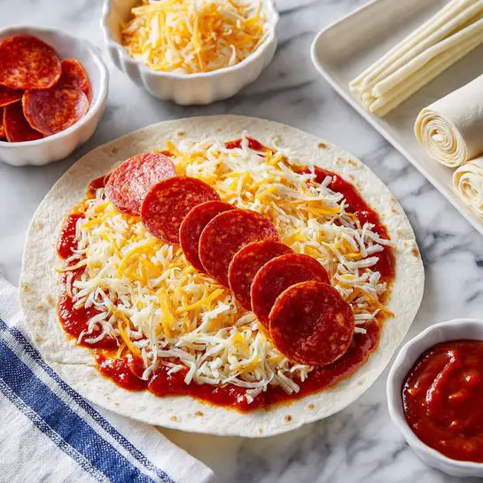 A round tortilla on a white marbled surface is layered with a bright red tomato sauce spread evenly over it, followed by a sprinkling of shredded yellow and white cheese scattered across the sauce. Near the center, a row of round, red pepperoni slices is placed horizontally, with a rolled stick of white string cheese laid right below them. In the background, three small white bowls hold extra ingredients: a scalloped bowl with mixed shredded cheese, a heart-shaped bowl filled with red tomato sauce, and a square-shaped bowl containing pepperoni slices. A larger white scalloped bowl also holds a stick of string cheese, and a white cloth with blue stripes rests nearby. photo taken with an iphone --ar 4:5 --v 7