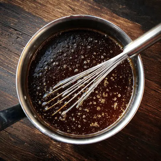 The image shows a close-up view of several small nut tarts in a metal muffin tray. Each tart has one layer of golden, textured pastry forming the base and sides, holding a thick layer of sticky, glossy caramel filling mixed with whole walnut pieces on top. One tart is being lifted by a woman's hand, prominently showing the shiny, brown caramel and crunchy walnuts that fill the tart, with the pastry edges slightly crisp and browned. The white marbled surface reflects softly in the background. photo taken with an iphone --ar 4:5 --v 7