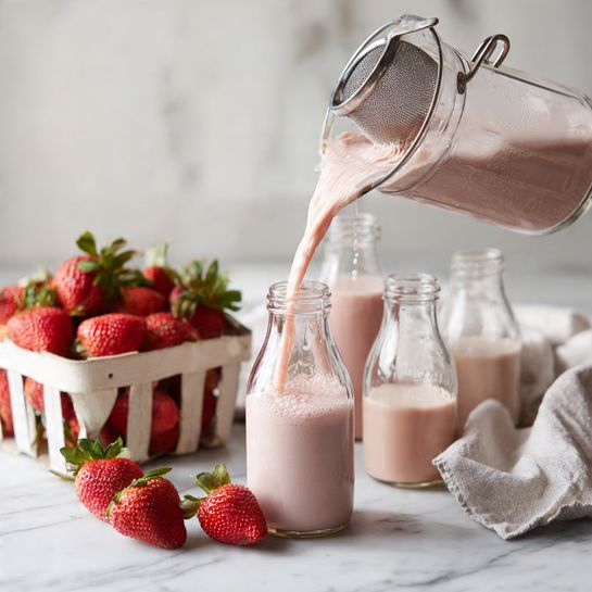 Six small clear jars filled with light pink creamy smoothies are placed on a white marbled surface. Each jar has a black lid tightly closed on top. In the background, there is a white square bowl filled with fresh bright red strawberries, and a white cloth with gray stripes is partially visible behind the bowl. The setting is clean and bright with soft natural light. photo taken with an iphone --ar 4:5 --v 7