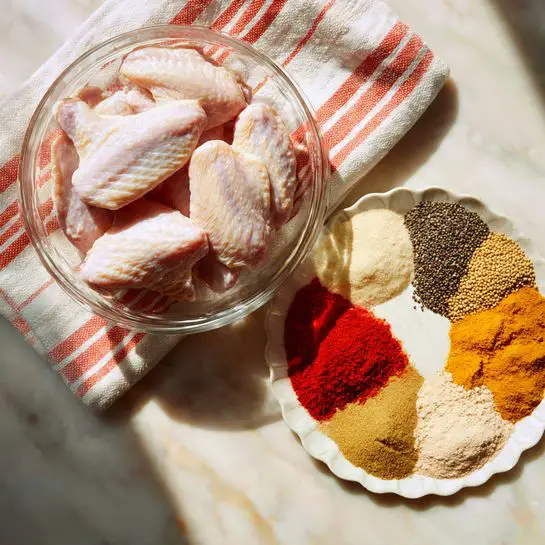 The image shows a clear glass bowl filled with many raw chicken wings that have a pale pink color and some smooth skin texture. The bowl is lined with a striped kitchen towel inside. Above the bowl, a white, scallop-edged plate holds six different spices, each in separate sections: a coarse black and white pepper mix, a pale yellow seasoning, a light brown powder, a bright orange-red powder, an off-white powder, and a beige powder, all arranged like slices of a pie. The background surface is white with a light marbled texture, and soft sunlight highlights the scene. Photo taken with an iphone --ar 4:5 --v 7