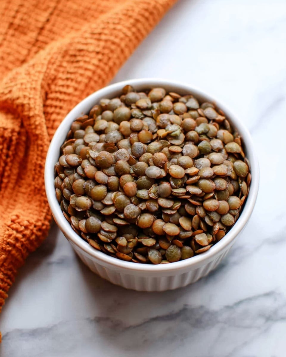 A small white ramekin cup is filled to the top with cooked lentils, showing their round, flat shapes in shades of brown and green. The lentils have a soft, slightly shiny texture. The ramekin sits on a white marbled surface with an orange textured cloth partially visible in the background on the left side. photo taken with an iphone --ar 4:5 --v 7