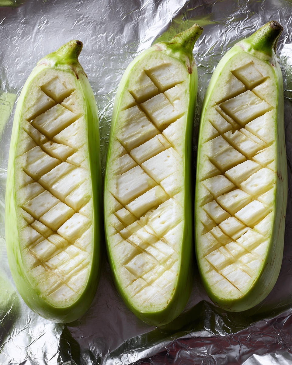 Three long light green eggplants are shown lying cut in half lengthwise on a silver foil surface. Each eggplant half has shallow diagonal cuts creating a grid pattern on the pale inside flesh, with the outer skin forming a thin green border. The silver foil underneath reflects light and has a slightly crumpled texture. photo taken with an iphone --ar 4:5 --v 7