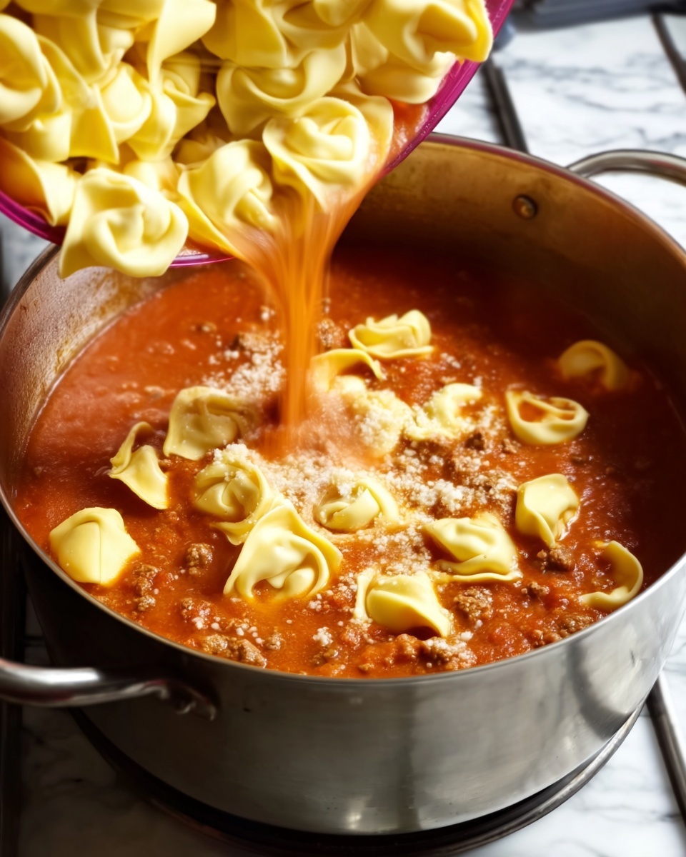 The image shows a white square bowl filled with a saucy tortellini dish. The dish has about three main layers: the bottom layer is a red tomato broth with visible pieces of diced tomatoes, the middle layer has browned ground meat mixed throughout, and the top layer features yellow tortellini pasta floating on top. The tortellini has a smooth texture and is sprinkled with small green parsley leaves. The dish looks warm and rich, with the broth slightly shiny and oily on the surface. The bowl is placed on a soft, white waffle-textured cloth, and all of this rests on a white marbled surface. Photo taken with an iphone --ar 4:5 --v 7