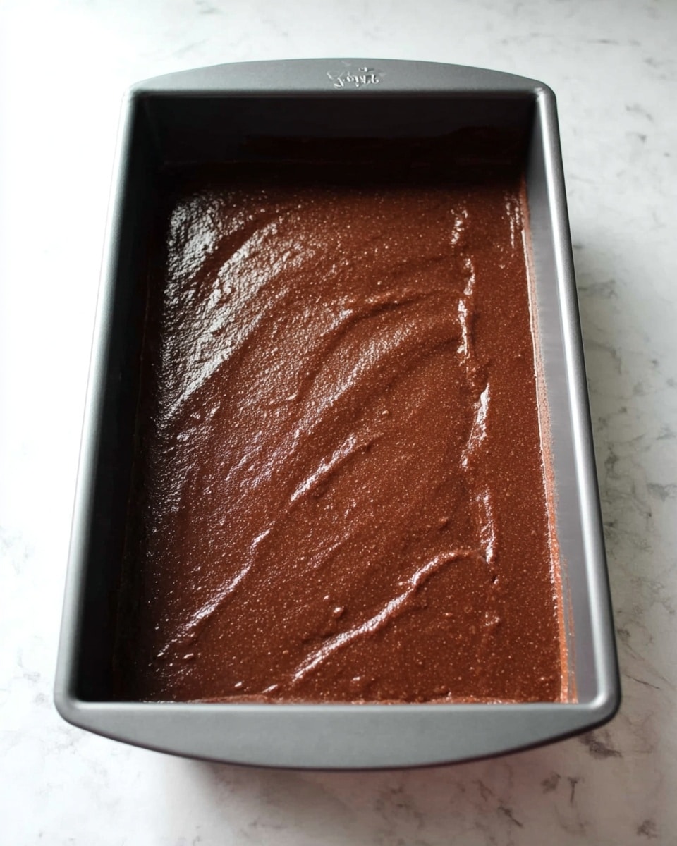 A dark brown chocolate batter fills a gray metal rectangular baking pan fully and is spread smoothly with some uneven texture on the surface. The pan is placed on a white marbled texture surface. Photo taken with an iphone --ar 4:5 --v 7