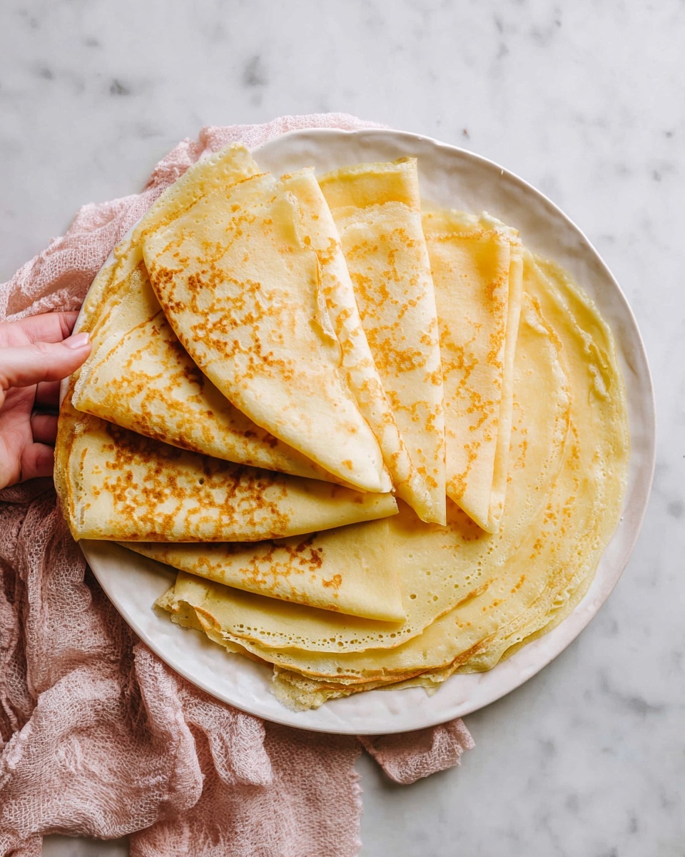 The image shows a simple meal setup centered around a single round pale yellow crepe with light brown spots on a white speckled plate. To the left, another white plate holds multiple folded crepes with similar coloring, arranged in a fan-like pattern. Near the top middle, there is a small glass of bright orange juice next to a jar of honey with a wooden honey dipper inside. Below that, a small round dish holds dark purple jam with a small black spoon resting on the edge. On the right side, a white plate with a few folded crepes is set with a gold fork and knife. Bottom left corner shows a small white bowl filled with mixed blackberries and raspberries. Additional elements include two more glasses of orange juice and a small yellow bowl with rich dark brown chocolate spread and a brass spoon. Everything is placed on a white marbled surface with a soft pink cloth partially visible at the bottom right. photo taken with an iphone --ar 4:5 --v 7