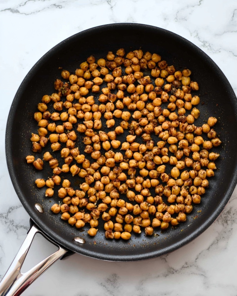 A black frying pan with a silver handle is filled with small, round, golden-brown chickpeas that have a slightly crispy texture, spread evenly across the pan's surface. The pan is placed on a white marbled surface, and the chickpeas show variations in color from light golden to a deeper brown, indicating they are cooked and roasted. The background is bright and clean, emphasizing the warm tones of the chickpeas. photo taken with an iphone --ar 4:5 --v 7