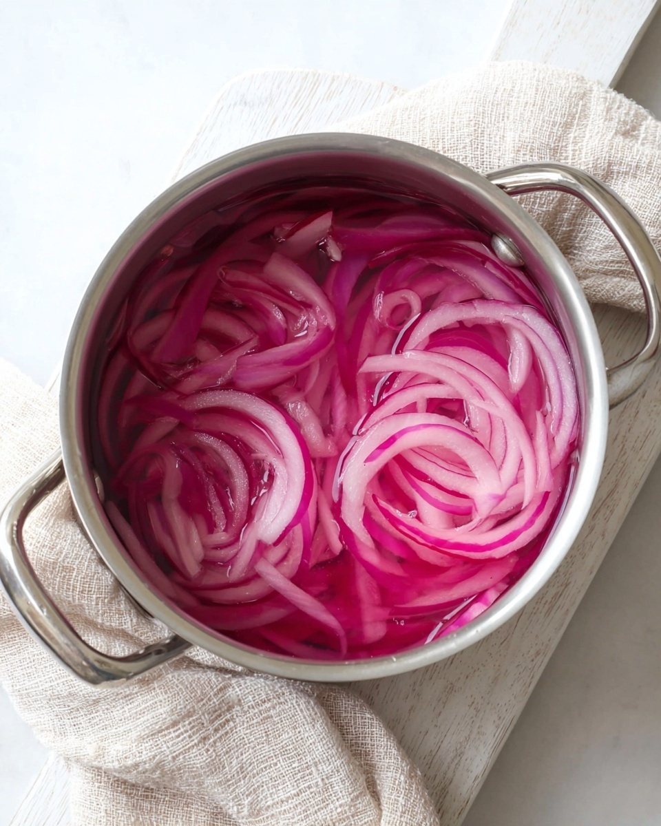 A shiny silver pot is filled with thin slices of bright pink and light purple onions soaking in clear, slightly pinkish liquid. The onions are layered loosely inside, showing their curved, translucent texture. The pot is placed on a white wooden board, and a light beige textured cloth is loosely folded in the background on a white marbled surface. The overall look is fresh and simple. photo taken with an iphone --ar 4:5 --v 7