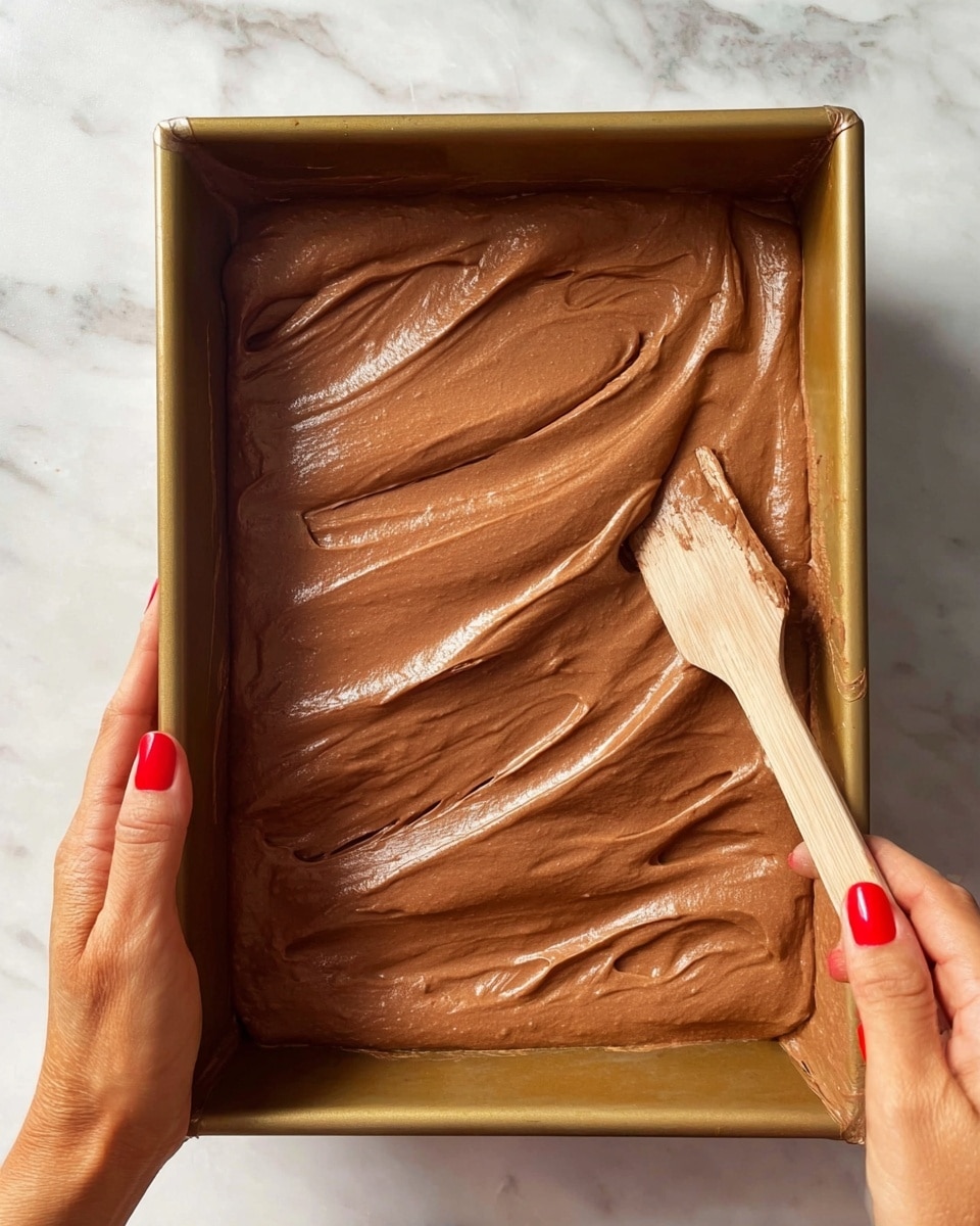 A rectangular baking pan is shown filled with one thick layer of smooth, light brown chocolate batter being spread evenly with a wooden spatula by a woman's hand with red nail polish, while another woman's hand with red nails holds the pan steady on a white marbled surface; the batter has a creamy texture with visible gentle waves from spreading, covering the entire base of the pan photo taken with an iphone --ar 4:5 --v 7