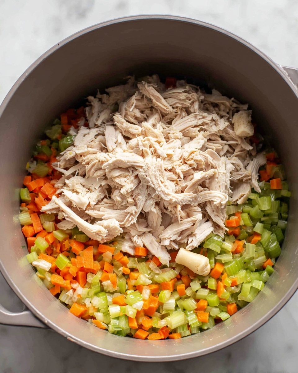 A close-up view inside a gray cooking pot shows a layer of finely chopped vegetables at the bottom, with bright orange carrots and green celery mixed with small bits of white onion, all chopped small and evenly spread out. On top of the vegetables is a pile of shredded light beige cooked chicken, with tender, stringy pieces layered loosely. A single piece of light brown bone sits near the center above the shredded chicken. The pot rests on a white marbled surface. photo taken with an iphone --ar 4:5 --v 7