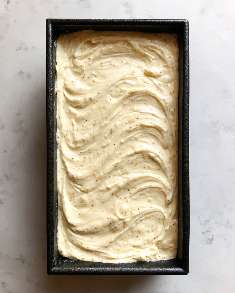 A rectangular black-edged tray filled with a thick, creamy mixture spread evenly across the surface. The mixture is light beige with small darker specks spread throughout, creating a slightly textured look. The top is smooth with soft swirls and gentle peaks, giving it a rich and fluffy appearance. The tray sits on a white marbled textured surface. photo taken with an iphone --ar 4:5 --v 7