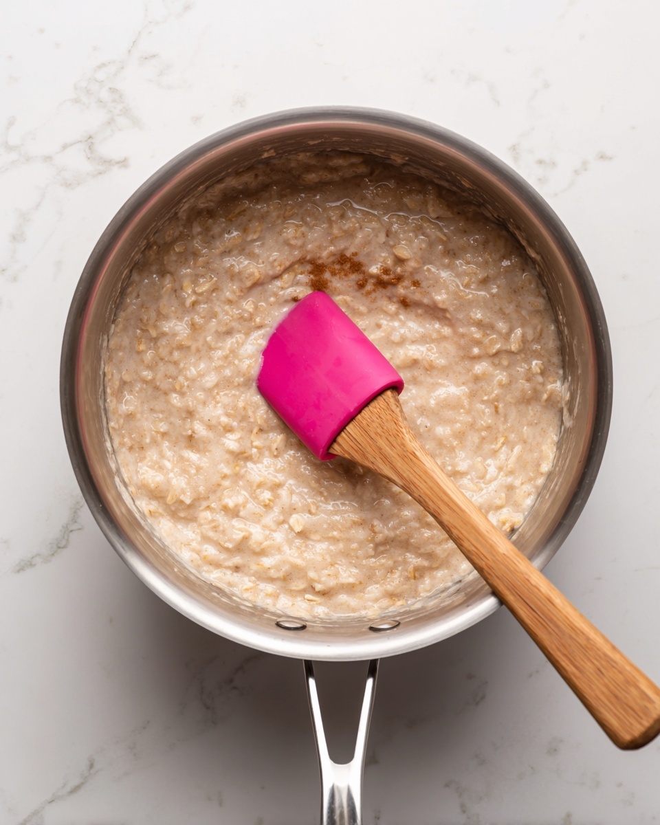 Inside a shiny metal pot, there is a thick layer of creamy oatmeal mixed with a cinnamon stick visible near the center. A wooden spatula with a bright pink silicone head rests in the oatmeal on the right side. The oatmeal is beige with a soft, textured appearance, filling almost the entire pot. The pot is open, showing its shiny metal rim and smooth inner surface. The scene is set on a white marbled surface. photo taken with an iphone --ar 4:5 --v 7