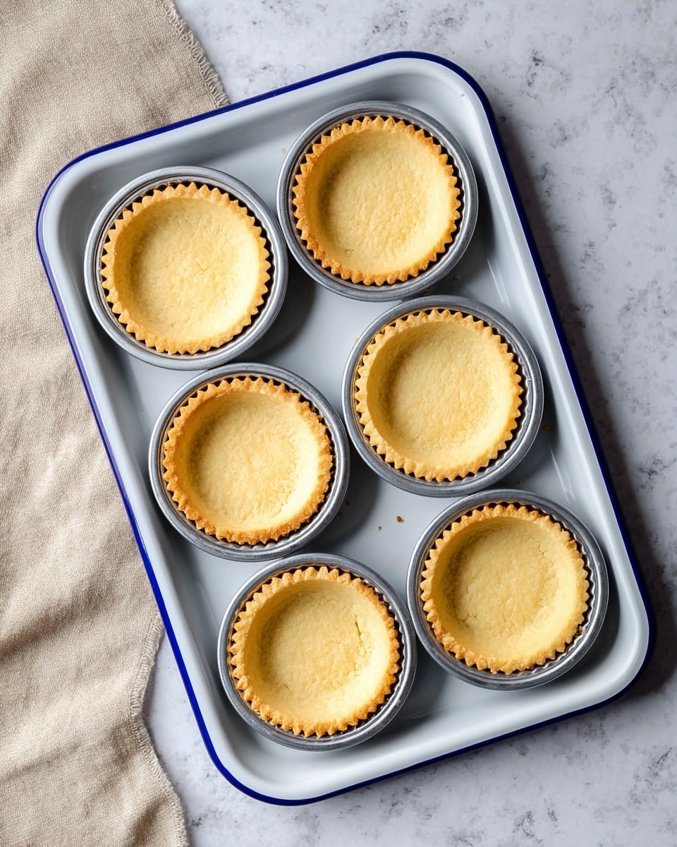 The image shows six empty tart shells inside shiny silver round molds on a white baking tray with a blue rim. Each tart shell has a golden brown, slightly crumbly crust with scalloped edges and a small hole in the center of the base. The baking tray is placed on a white marbled surface with a beige cloth partially under it on the left side. The lighting highlights the smooth texture of the tart shells and the clean lines of the tray photo taken with an iphone --ar 4:5 --v 7