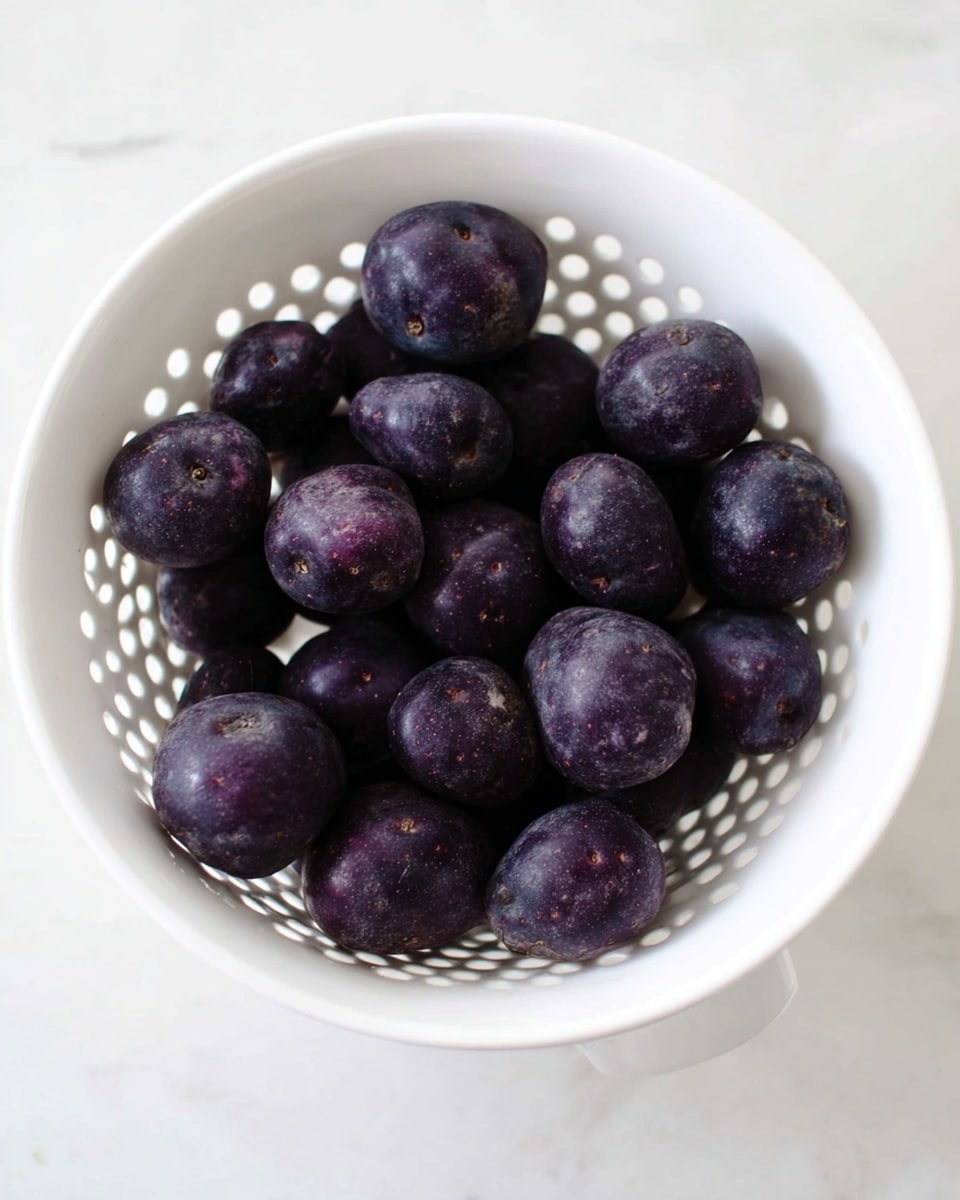 A close-up view of a single purple potato rose inside a round metal muffin tin cup, made from thin slices of deep purple potato arranged in layered, curled petals to resemble a flower, with a slightly rough texture and light seasoning specks on the surface. The background shows other similar purple potato roses in the muffin tin, all placed on a white marbled surface. The lighting is soft and natural, highlighting the rich purple shades and subtle textures of the potato petals photo taken with an iphone --ar 4:5 --v 7