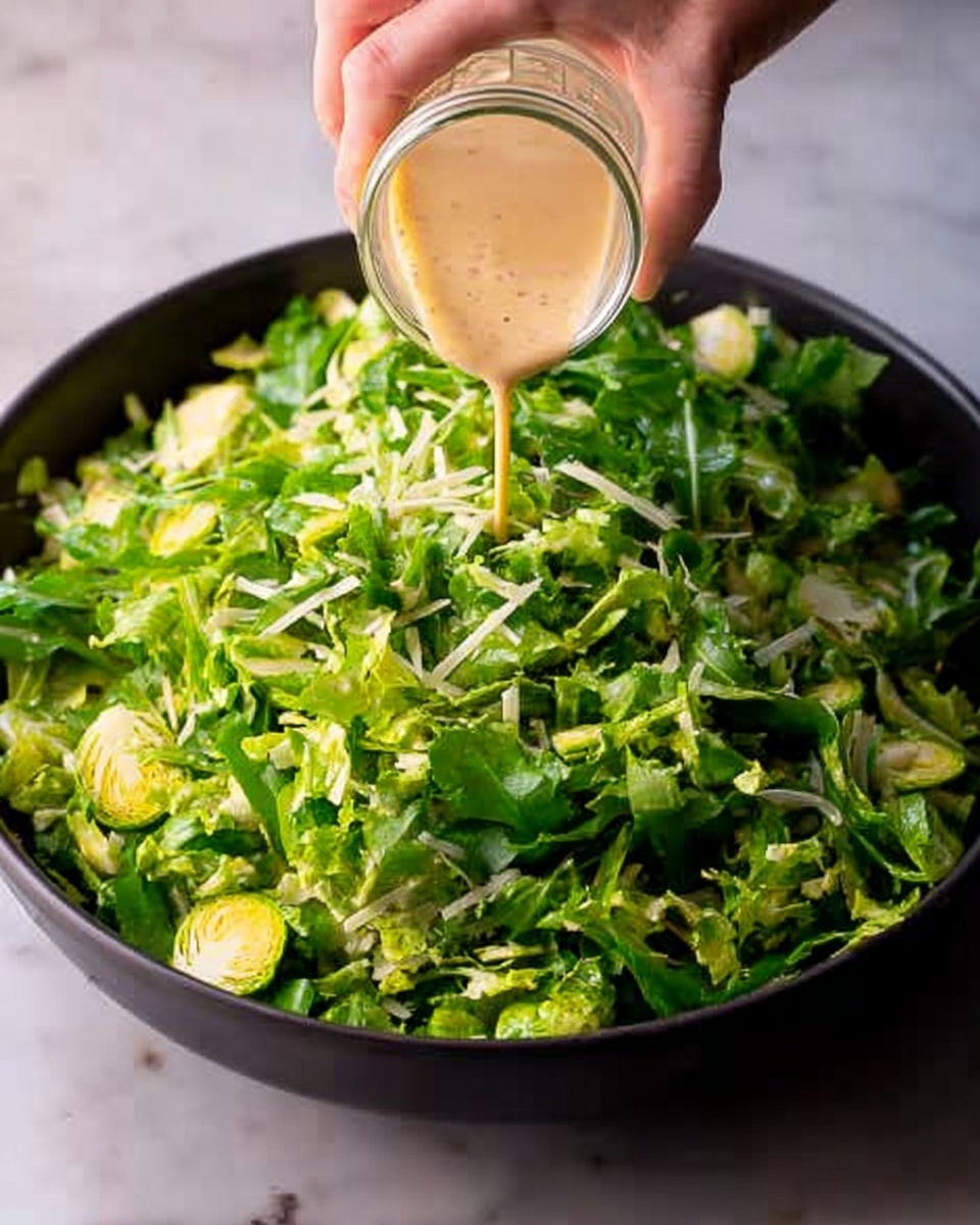 A close-up of a black bowl filled with fresh green salad leaves, including arugula and Brussels sprouts pieces, all mixed and piled evenly. A woman's hand is pouring a creamy light brown dressing from a clear glass jar over the salad. The bowl sits on a white marbled surface, and the lighting highlights the fresh texture of the greens and the smooth flow of the dressing. photo taken with an iphone --ar 4:5 --v 7