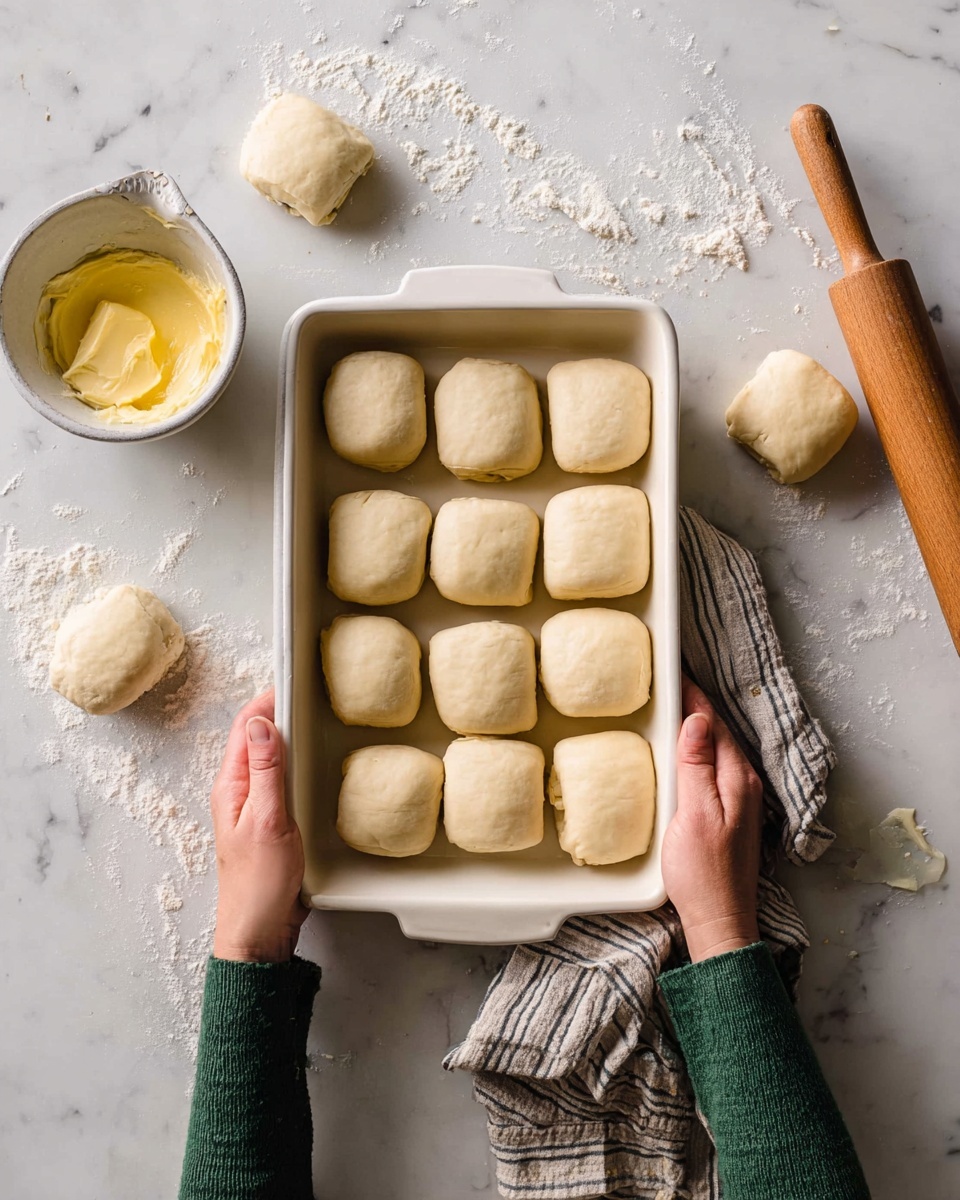 The image shows a white rectangular baking tray filled with ten small rolled dough parcels arranged in two uneven rows inside the tray. The dough parcels are pale beige and smooth, with a soft texture. Around the tray on a white marbled surface, there are four more rolled dough pieces scattered, a wooden rolling pin with a light brown color, and a small white bowl filled with a yellow butter or spread, with a spreader knife resting inside. Two woman’s hands wearing green sleeves hold the baking tray from the left and right sides, with a striped cloth partially visible near the right hand. There is some flour scattered on the marbled surface, adding a rustic kitchen feel. photo taken with an iphone --ar 4:5 --v 7