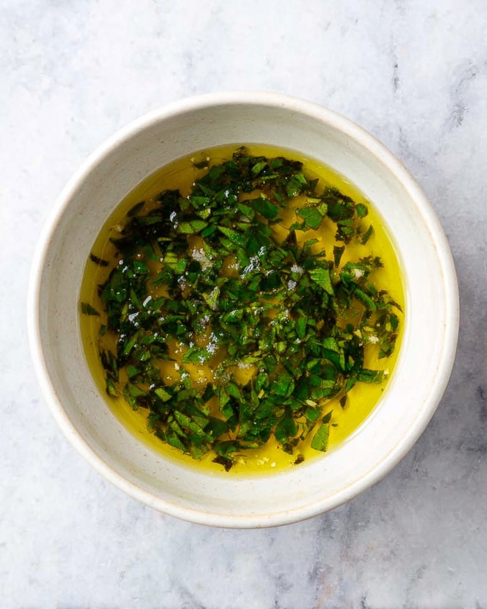 A white bowl sits on a white marbled surface, filled with a greenish-yellow oily mixture with small pieces of dark green chopped herbs evenly spread throughout. The mixture has a slightly shiny texture, with some visible small granules of salt or seasoning on top. The bowl has smooth edges and is viewed from above, showing the ingredients clearly inside. photo taken with an iphone --ar 4:5 --v 7