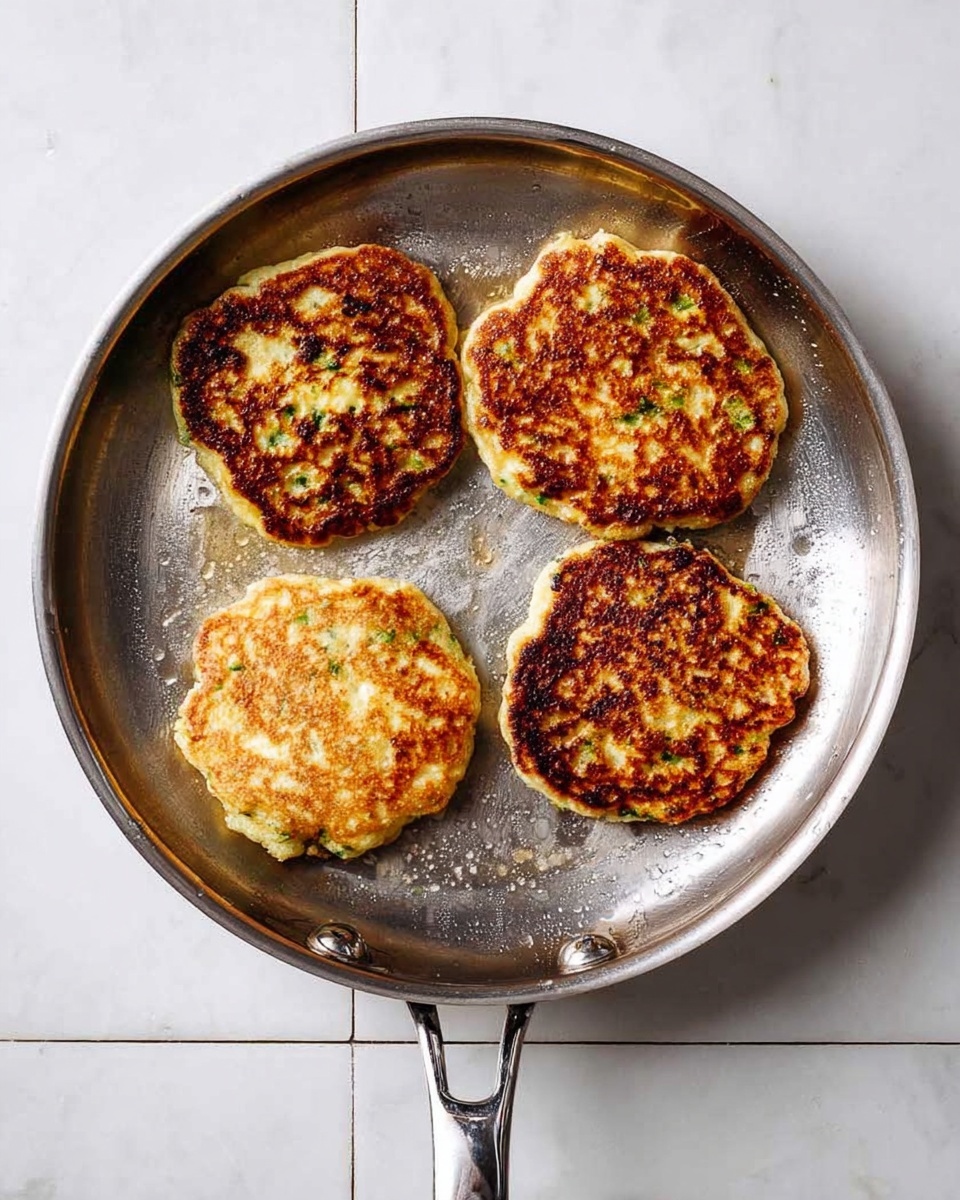 A silver frying pan with a shiny surface holds four round, golden-brown pancakes that are slightly uneven in shape. The pancakes have a mix of light and dark brown spots with green bits visible, giving texture and color to the surface. The pan rests on a white marbled tile background, and the handles of the pan are visible at the top and bottom edges. photo taken with an iphone --ar 4:5 --v 7