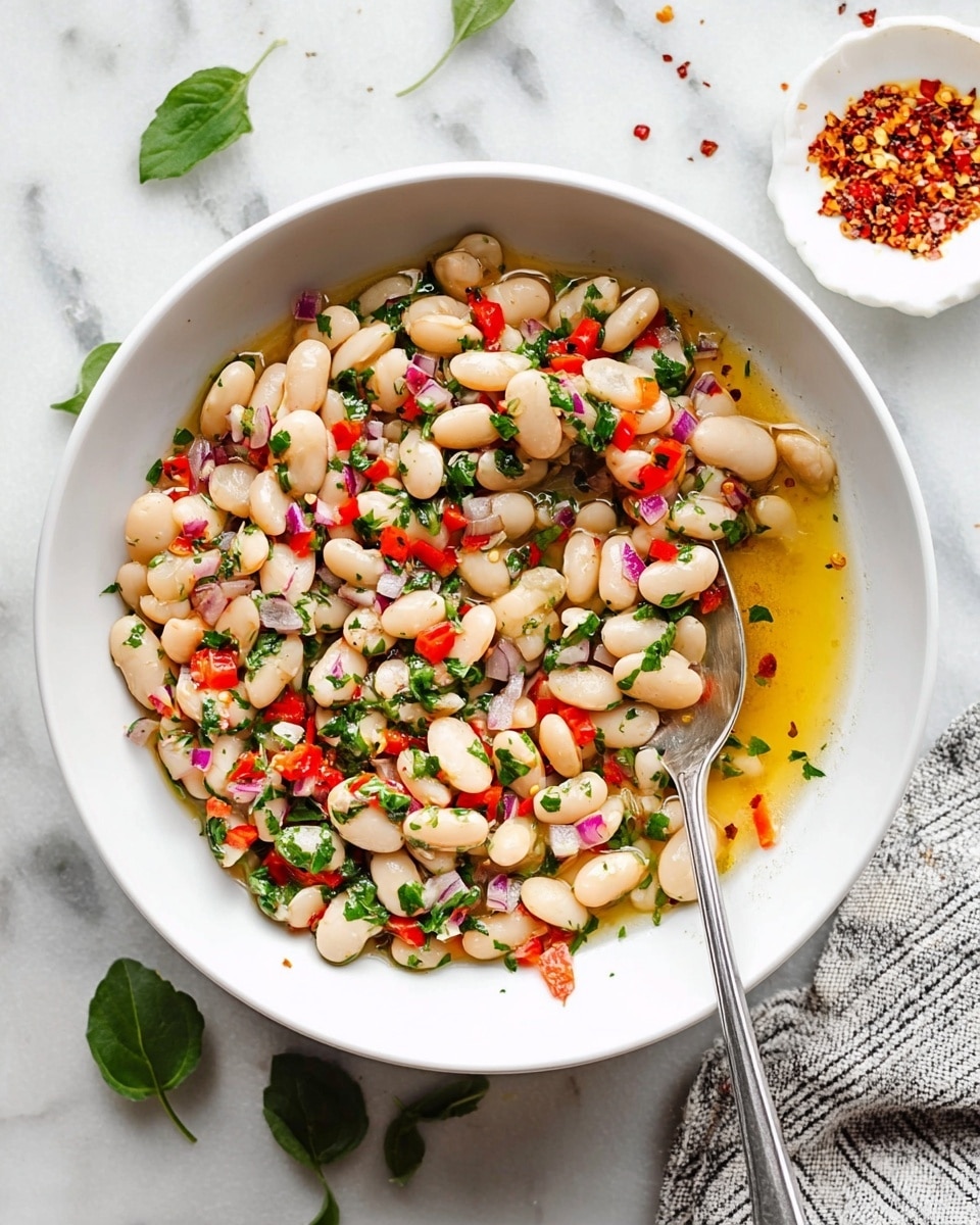 A white bowl filled with a colorful bean salad showing one layer of white beans mixed evenly with small pieces of red bell pepper, finely chopped red onion, and bright green herbs, all coated lightly in a golden liquid dressing that pools slightly in the center. A silver spoon rests inside, scooping up some of the salad, and the bowl is placed on a white marbled surface with scattered green leaves nearby and a small white dish with red chili flakes on the top right corner. A white and black striped cloth is partially visible at the bottom right. Photo taken with an iphone --ar 4:5 --v 7