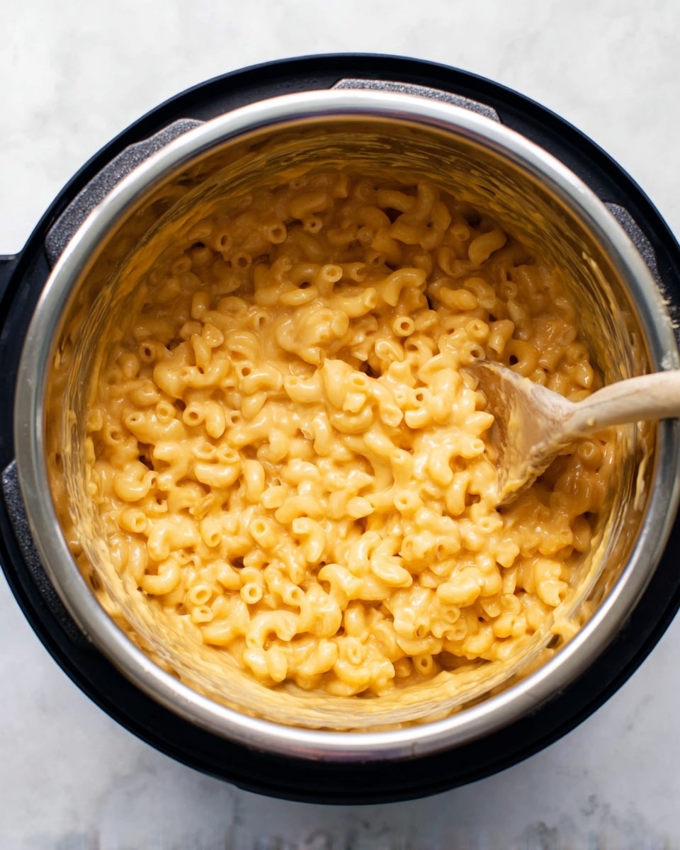 The image shows a close-up of creamy macaroni and cheese inside a shiny silver pot with a black rim, sitting on a white marbled surface. The macaroni is covered in a smooth, light orange cheese sauce, with a thick texture that clings to the small, curved pasta pieces. A wooden spoon with cheese sauce on it rests inside the pot, partially submerged in the macaroni, suggesting the dish is freshly stirred. The photo is taken from directly above, capturing the full round shape of the pot and the rich, cheesy pasta inside, photo taken with an iphone --ar 4:5 --v 7