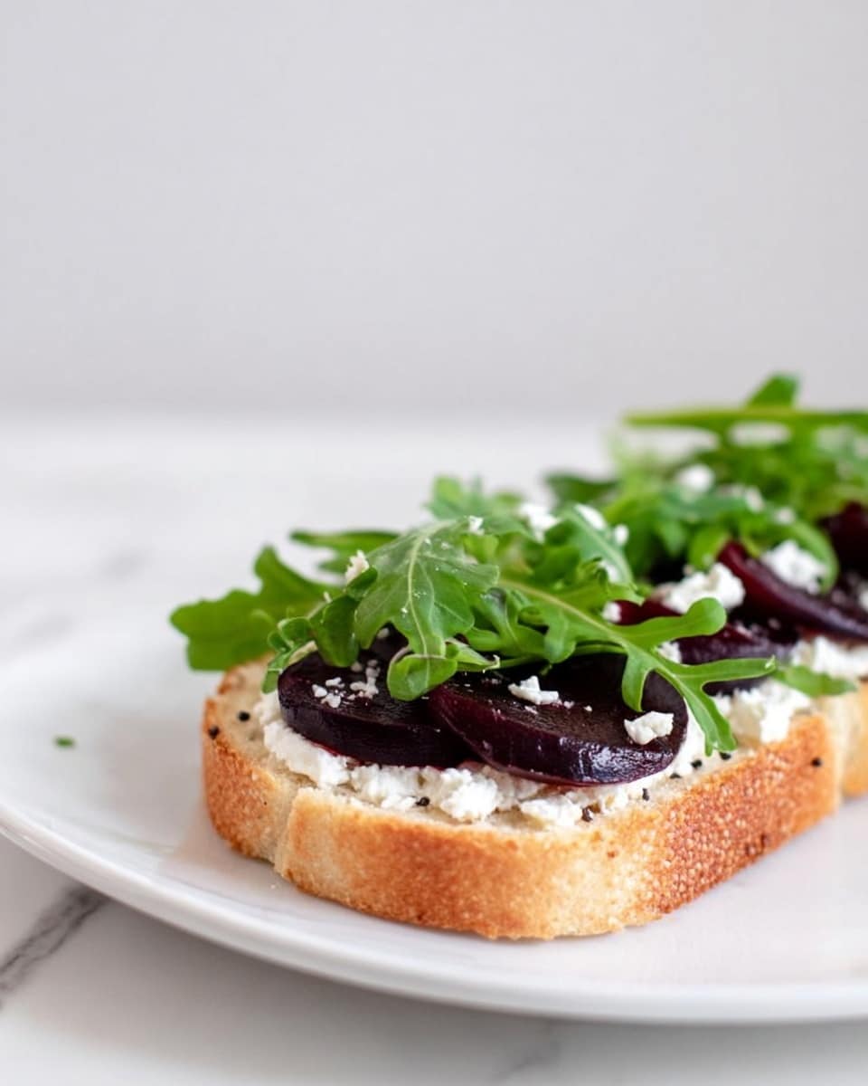 A slice of toasted bread serves as the base, topped with a layer of white creamy cheese spread evenly. On top of the cheese, there are several thin, dark purple slices of beet arranged in a single layer. The top layer is fresh green arugula leaves, scattered loosely. The toast sits on a white plate with a white marbled surface in the background. The photo is taken with an iphone --ar 4:5 --v 7