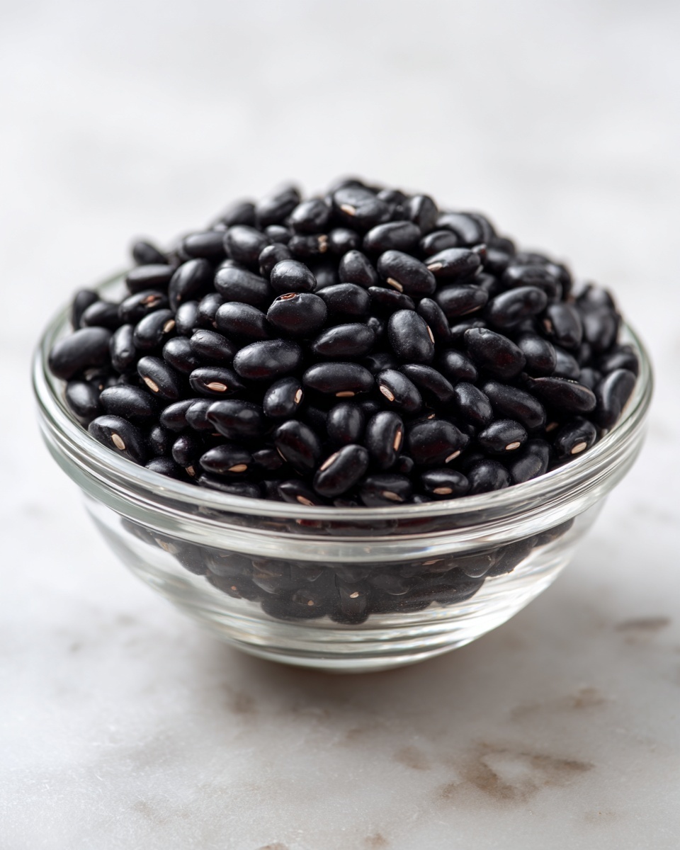The image shows a close-up of a clear glass bowl filled with small, shiny black beans that have a smooth texture. The bowl is placed on a white marbled surface, and the focus is on the front pile of beans, leaving the rest softly blurred in the background. There are no other layers or colors visible, just the solid black beans in the transparent bowl. photo taken with an iphone --ar 4:5 --v 7