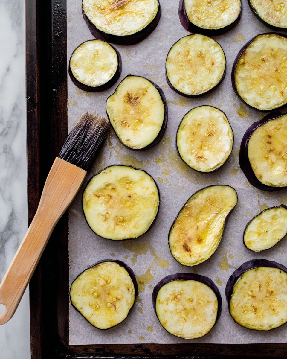 The image shows a baking tray lined with parchment paper holding several eggplant slices of different sizes spread out evenly. Each slice has a pale yellow interior with visible seeds and a dark purple skin around the edges. Some slices appear to be brushed with oil, giving them a shiny texture. A wooden brush with dark bristles rests on one of the eggplant slices on the left side. The tray edges are dark, and the background is a white marbled texture. photo taken with an iphone --ar 4:5 --v 7