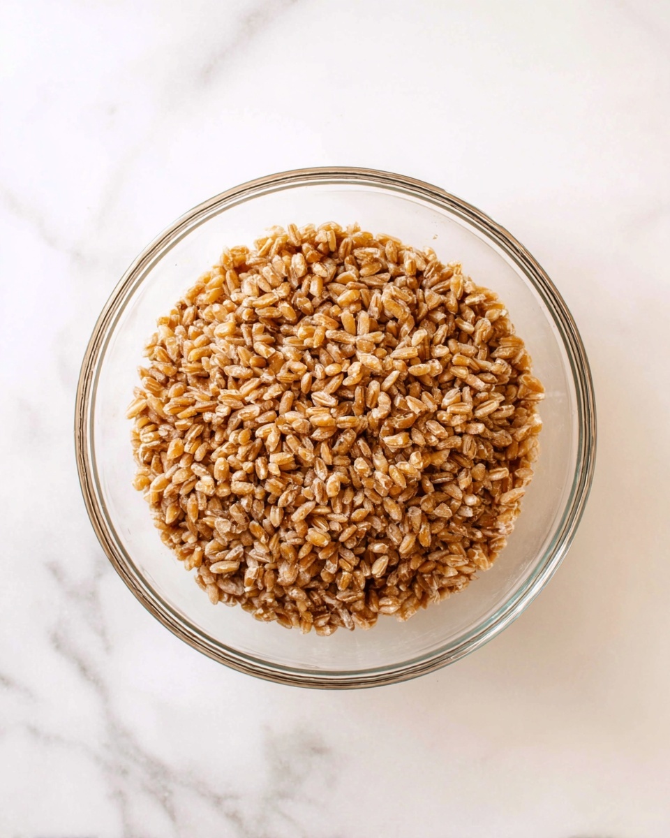 A clear glass bowl filled with cooked farro grains, showing a dense layer of firm, light brown, slightly shiny grains. The grains have a rough, chewy texture visible on the surface, tightly packed inside the bowl. The bowl sits on a white marbled surface with soft gray veins. The photo is taken from directly above, emphasizing the round shape of the bowl and the uniform texture of the farro. Photo taken with an iphone --ar 4:5 --v 7