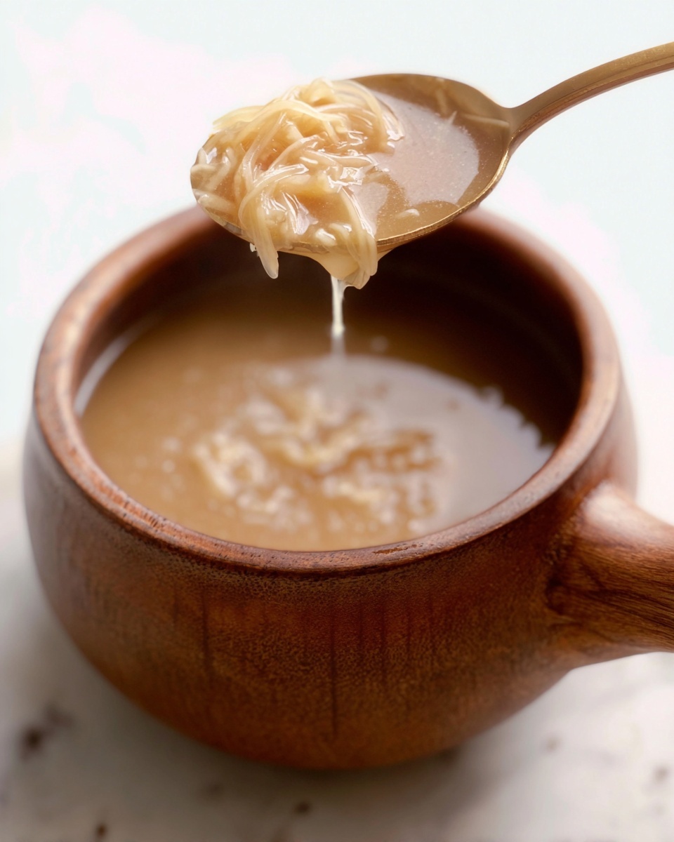 A close-up image shows a wooden cup filled with light brown soup that appears smooth with small chunky bits. Above the cup, a spoon holds a portion of the soup with translucent and slightly shiny thin noodle strands and soft small pieces, dripping back into the cup. The cup sits on a white marbled surface, with the wooden handle visible on the right side. The photo is softly lit with a shallow depth of field, focused on the soup in the spoon. Photo taken with an iphone --ar 4:5 --v 7