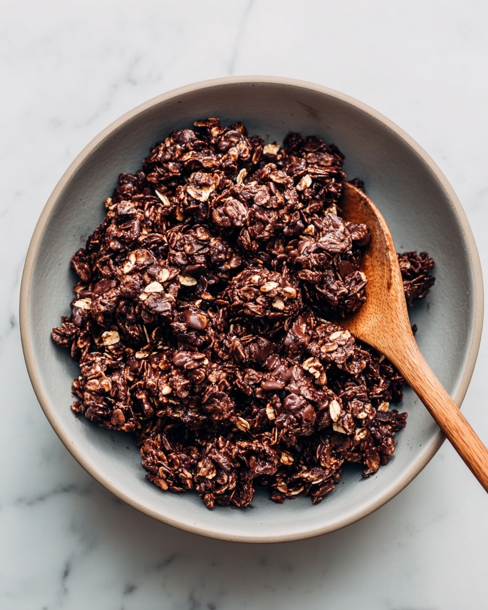 The image shows a close-up view of a bowl filled with thick clusters of dark brown chocolate oat granola mixed with visible oats. The texture looks rough and chunky with a mix of glossy and matte patches, showing the oats coated evenly in chocolate while some oats stick out more clearly. There is a wooden spoon partially buried in the granola on the right side, its spoon part covered in the mixture. The bowl is light grey and round, sitting on a white marbled surface. photo taken with an iphone --ar 4:5 --v 7