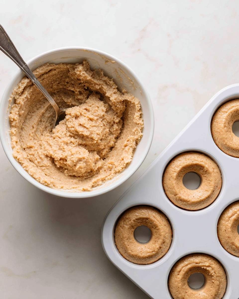 The image shows two close-up photos side by side on a white marbled surface. On the left, there is a rough, thick light brown batter with a grainy texture being stirred with a large silver spoon inside a bowl that is not fully visible. The batter looks soft and dense with visible tiny air pockets. On the right, there is a white muffin pan with four donut-shaped portions of the same light brown batter evenly spread inside the circular molds. The batter fills the molds to a medium height with smooth tops, and the empty central holes are clear and round. Photo taken with an iphone --ar 4:5 --v 7