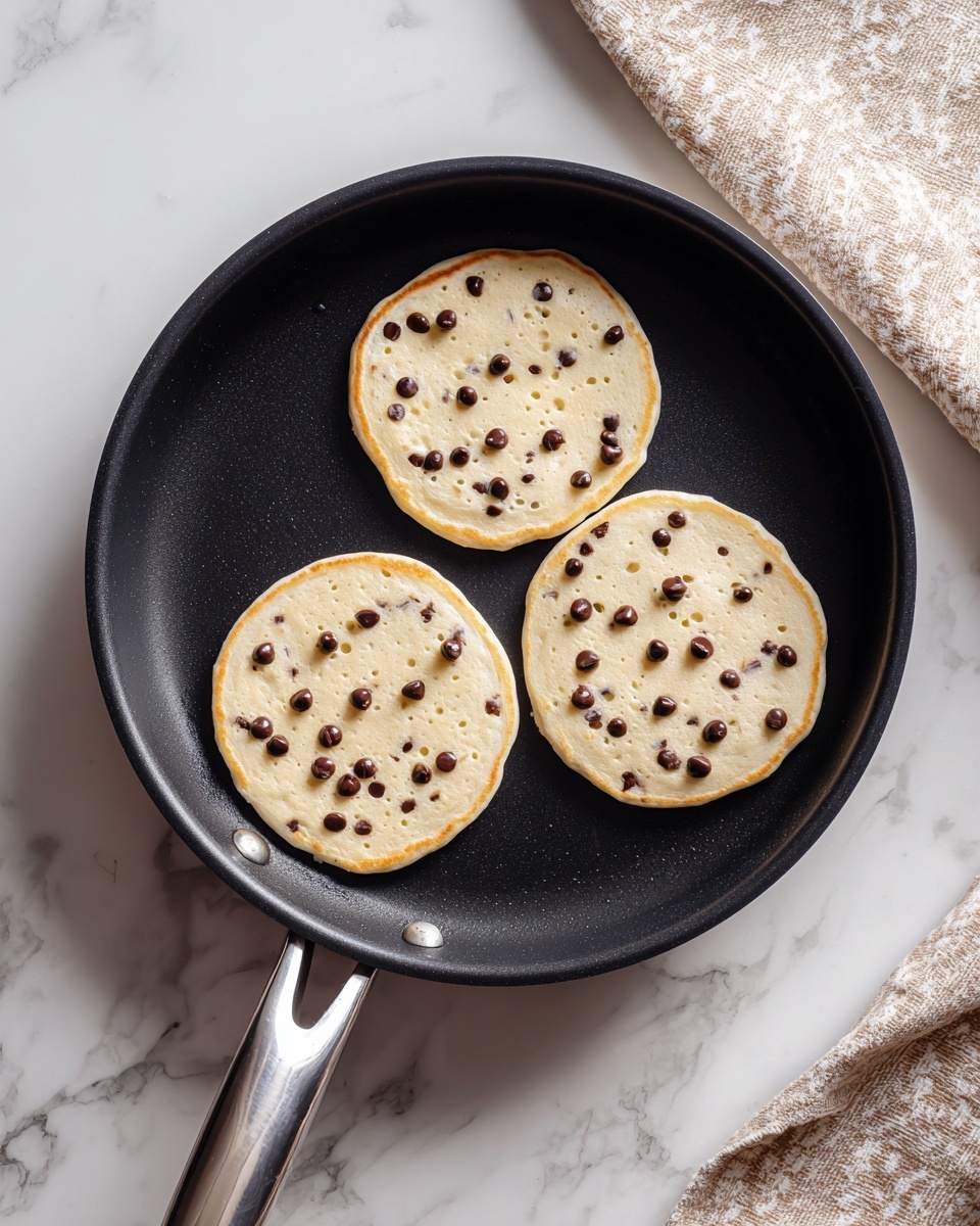Three raw pancakes with chocolate chips are cooking in a black frying pan. Each pancake is round and light beige with small dark chocolate chips spread evenly throughout the batter. The pancakes are arranged in a triangular pattern inside the pan, which has a silver handle. The pan sits on a surface with a white marbled texture, and a beige and white patterned cloth is partially visible in the top right corner. photo taken with an iphone --ar 4:5 --v 7