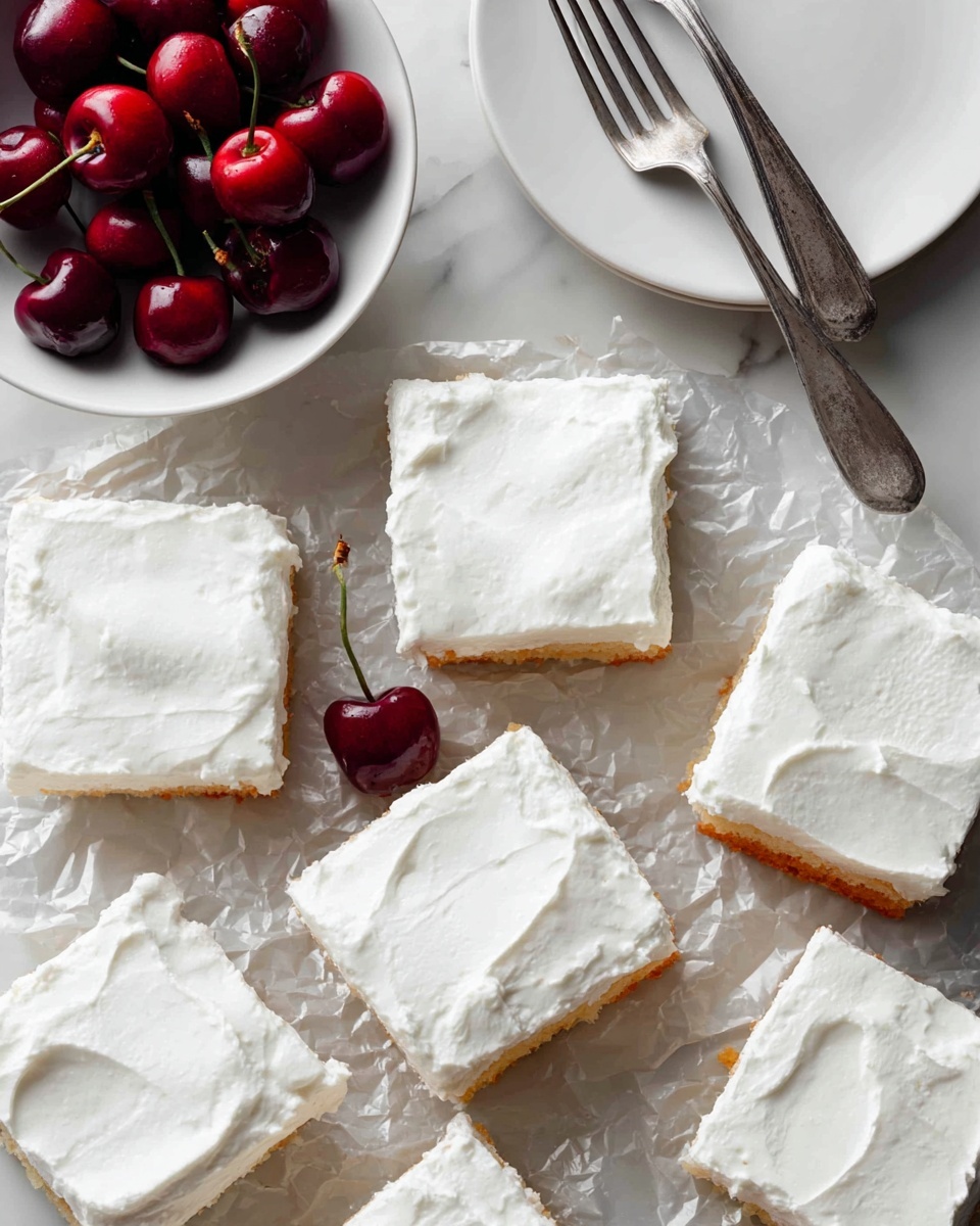 Nine square pieces of a dessert with a thick white frosting on top are placed on crinkled silver foil. The frosting layer is smooth but slightly textured, covering a light golden cake base that is visible along the edges. One piece in the top right corner is slightly moved out of alignment. Part of a white bowl with bright red cherries sits in the top left corner, and two metal forks rest on a white plate near the top right. The surface underneath is a white marbled texture photo taken with an iphone --ar 4:5 --v 7