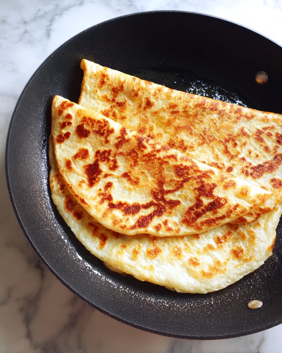 A golden brown folded flatbread with a crispy texture on top, showing light and darker brown spots, is cooking in a black pan with slight oil glistening around it. The flatbread looks soft near the edges, which are slightly puffed and pale, creating a contrast with the crispy center. The pan sits on a white marbled surface. Photo taken with an iphone --ar 4:5 --v 7