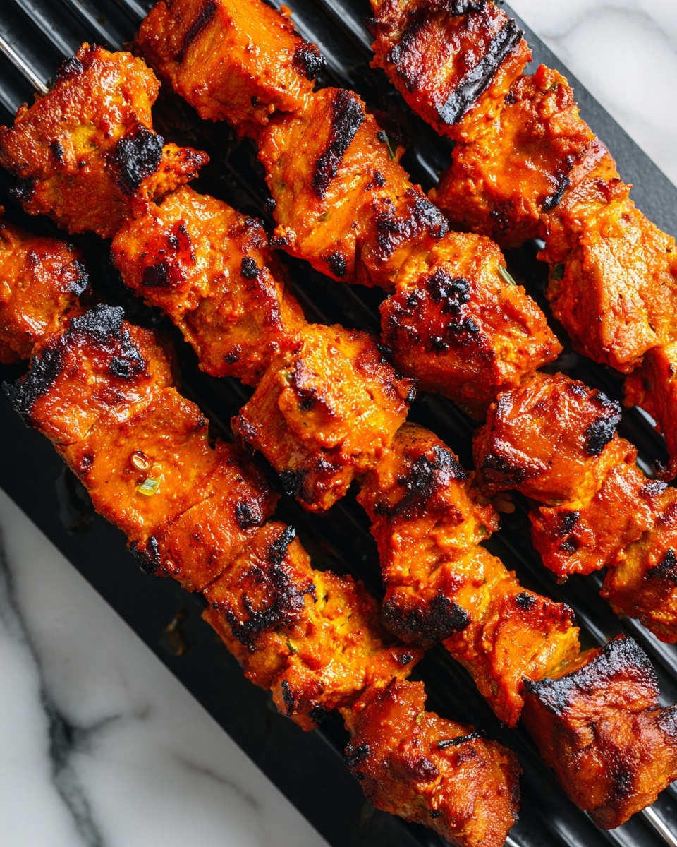 Four metal skewers, each holding several bright orange pieces of grilled meat, rest on a black grill surface. The meat pieces are thick and unevenly shaped, with some charred dark brown grill marks and a slightly crispy texture on the edges. The orange color suggests a rich marinade covering each piece, with a glossy, moist look. The skewers are placed close to each other, filling most of the frame, with the black grill lines visible between the meat chunks. The background is a white marbled texture. Photo taken with an iphone --ar 4:5 --v 7