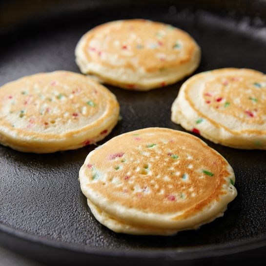 Cake Mix Pancakes Recipe 5 A stack of four thick, golden-brown pancakes with small colorful sprinkles cooked inside each layer sits on a white plate. On top, there is a dollop of white cream covered with bright rainbow sprinkles in pink, orange, green, blue, purple, and white. Around the plate and on the white marbled surface, more scattered colorful sprinkles add a fun and festive touch. The pancakes have a soft, slightly bubbling texture with edges that are a little crispy. The photo taken with an iphone --ar 4:5 --v 7