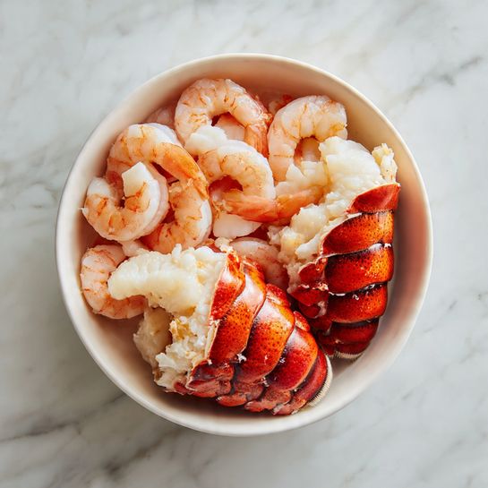 A white bowl on a white marbled surface filled with a mix of seafood. There are two large bright red lobster tails on one side, showing their segmented shells with some cracks revealing white meat inside. Surrounding the lobster tails, there is a layer of pink and white cooked shrimp arranged in a circular pattern, their curved shapes adding texture. The shrimp have a slightly translucent look, contrasting with the solid lashes of the lobster shells. photo taken with an iphone --ar 4:5 --v 7
