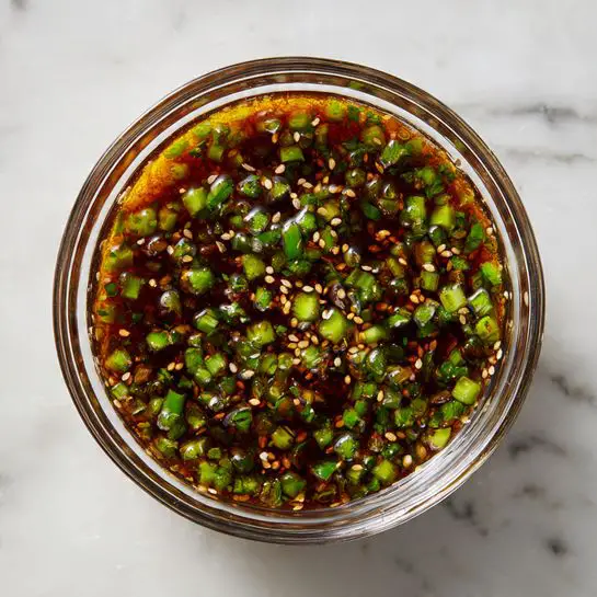 Sheet Pan Salmon with Garlic-Ginger Soy Glaze and Roasted Vegetables Recipe 4 A clear glass bowl filled with a dark brown sauce mixed with small green pieces of chopped herbs and scallions, plus many light tan sesame seeds scattered evenly throughout. The sauce has a slightly oily texture that reflects light, and the green bits add a fresh look contrasting with the rich brown liquid. The bowl sits on a white marbled surface. photo taken with an iphone --ar 4:5 --v 7