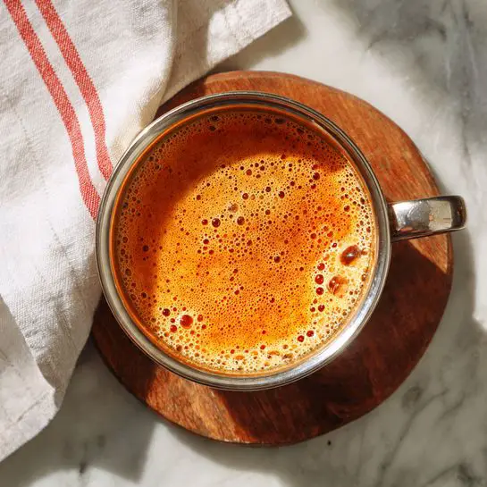 The image shows a close-up of a clear blender cup filled with a thick, bright orange mixture that has a smooth but slightly frothy texture near the top. The blender cup has black measurement markings from 250 to 1800 ml on the clear surface. The blender base is visible in a soft beige color. In the blurry white marbled background, there is a small white container and a hint of a red and white cloth. photo taken with an iphone --ar 4:5 --v 7