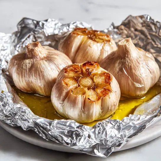 The image shows a close-up of roasted garlic bulbs wrapped partially in crinkled silver foil, with a visible layer of golden olive oil pooled at the bottom of the foil. The garlic cloves are exposed at the top, displaying a smooth, soft texture with a warm golden-brown color. The outer skin of the garlic cloves is light beige and slightly translucent, curling back around the edges. The background and surface is a white marbled texture. photo taken with an iphone --ar 4:5 --v 7