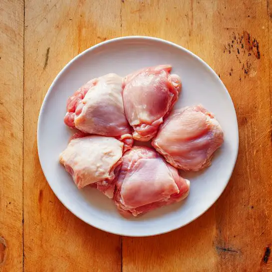 A white round plate holds six raw pieces of chicken thighs arranged in a slightly overlapping way, showing smooth, shiny pinkish flesh with patches of white fat along the edges. The plate is placed on a light brown wooden surface with natural grain and marks visible. The chicken pieces vary in size and shape but all have a moist texture. photo taken with an iphone --ar 4:5 --v 7