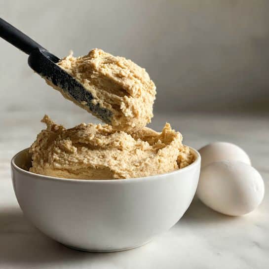The image shows four round cookies on a piece of brown parchment paper, which is placed on a black metal baking tray. The cookies have a light brown color with a slightly cracked surface texture, each having a circular shape with defined edges and a soft, slightly puffy appearance. The baking tray rests on a white marbled surface that adds a bright contrast to the warm tones of the cookies and the parchment paper. Photo taken with an iphone --ar 4:5 --v 7