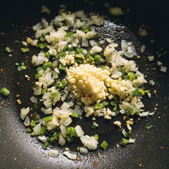 The image shows a close-up view of a dark pan with three layers of ingredients being cooked. The bottom layer consists of small white pieces of onion scattered across the pan, creating a soft and slightly translucent texture. Mixed with the onion are bits of green chili peppers adding a speckled pattern and a slightly glossy look. In the center, there is a small mound of light beige minced garlic that looks fresh and moist, standing out against the darker pan surface. The overall scene has a warm, cooking process feel and the pan’s black color contrasts with the light colors of the onions, chili peppers, and garlic. photo taken with an iphone --ar 4:5 --v 7
