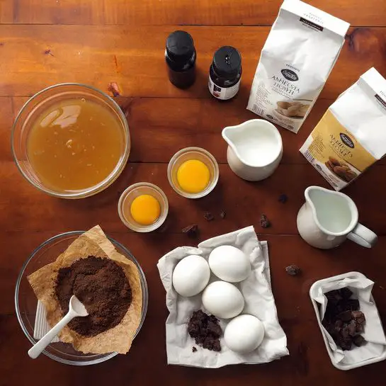 A dark brown round cake layer sits inside a metal round baking pan on a white marbled surface. A light cream-colored batter is being poured from a metal bowl over the cake layer, spreading smoothly across the top, creating a thick, soft layer. The batter looks creamy and slightly thick as it covers the darker cake base evenly. No other items or decorations are visible. photo taken with an iphone --ar 4:5 --v 7