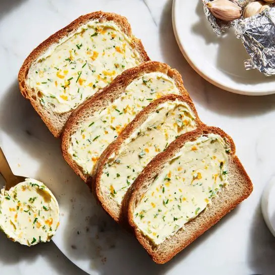 Four slices of bread lie on a white marbled surface, each spread with a thick layer of light yellow butter mixed with small green herbs and tiny orange bits. The butter is smooth and evenly spread, covering most of the top surface of each slice. To the side, there is a white plate with more butter and a spreader. In the top right corner, some foil-wrapped roasted mushrooms add a touch of brown and silver to the scene. The image looks simple and fresh. photo taken with an iphone --ar 4:5 --v 7