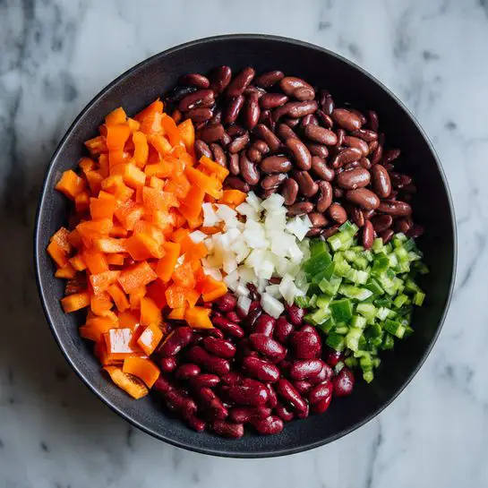 The image shows a black bowl filled with different chopped and whole ingredients placed in layers. At the bottom left, there are small bright orange bell pepper cubes. Next to these are many shiny dark brown beans spread across the left and center part of the bowl. To the right of the beans, there are bright red kidney beans covering the middle-right side. Behind the red kidney beans, there are small white garlic pieces and in front of that on the right side of the bowl, there are finely chopped green bell peppers. The bowl is placed on a white marbled surface. photo taken with an iphone --ar 4:5 --v 7