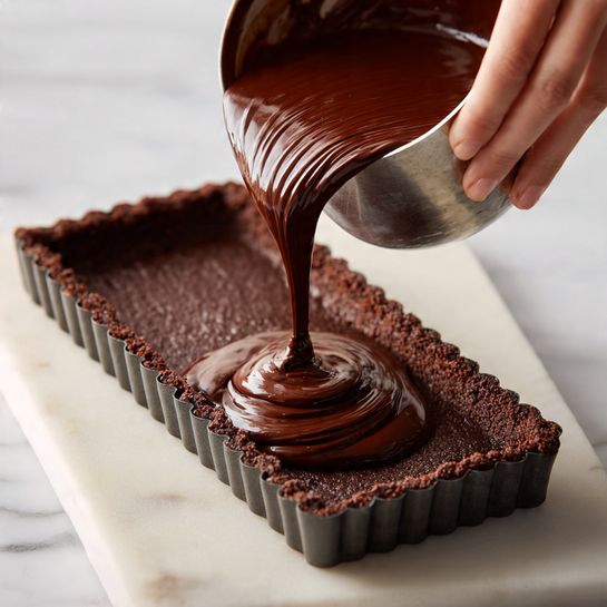 A long rectangular tart with scalloped edges is shown on a white marbled surface. The tart crust is dark brown and crumbly, evenly pressed inside the metal tart mold. A woman's hand is pouring thick, smooth dark chocolate filling from a silver mixing bowl onto the base layer inside the tart mold. The chocolate filling cascades in a rich, glossy layer over the crust, mostly in the center, waiting to spread. The scene captures the process of filling the tart crust with chocolate batter. Photo taken with an iphone --ar 4:5 --v 7