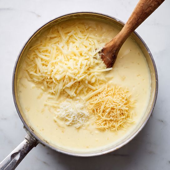 A close-up top view of a creamy cheese sauce in a pot. The sauce is smooth and pale yellow, covering the entire pot surface. On top, there are two piles of shredded pale yellow cheese, one on the right side and one smaller on the left, starting to melt into the sauce. A wooden spoon is visible at the top left edge, stirring the sauce gently. The pot is placed on a white marbled surface. photo taken with an iphone --ar 4:5 --v 7