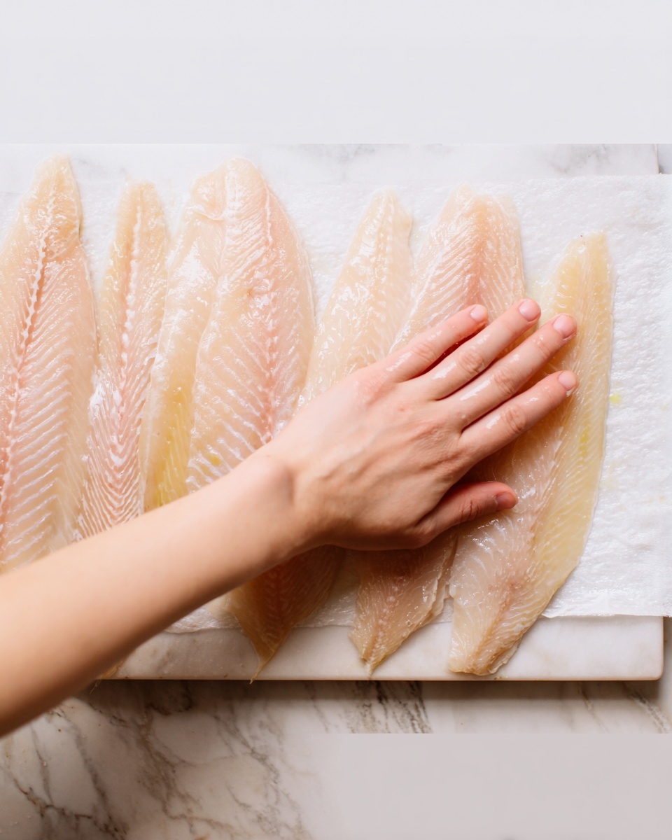 The image shows a white scalloped plate with two main layers arranged side by side: a golden-brown cooked chicken fillet sprinkled with chopped green herbs on the left, and a heap of fluffy white rice on the right. A bright yellow lemon half rests on the top right edge of the rice. On the plate’s left side, a silver fork is partially inserted into the rice near the chicken. The plate sits on a white marbled surface with a folded white cloth featuring thin black stripes to the right of the plate. The lighting is bright and natural. Photo taken with an iphone --ar 4:5 --v 7